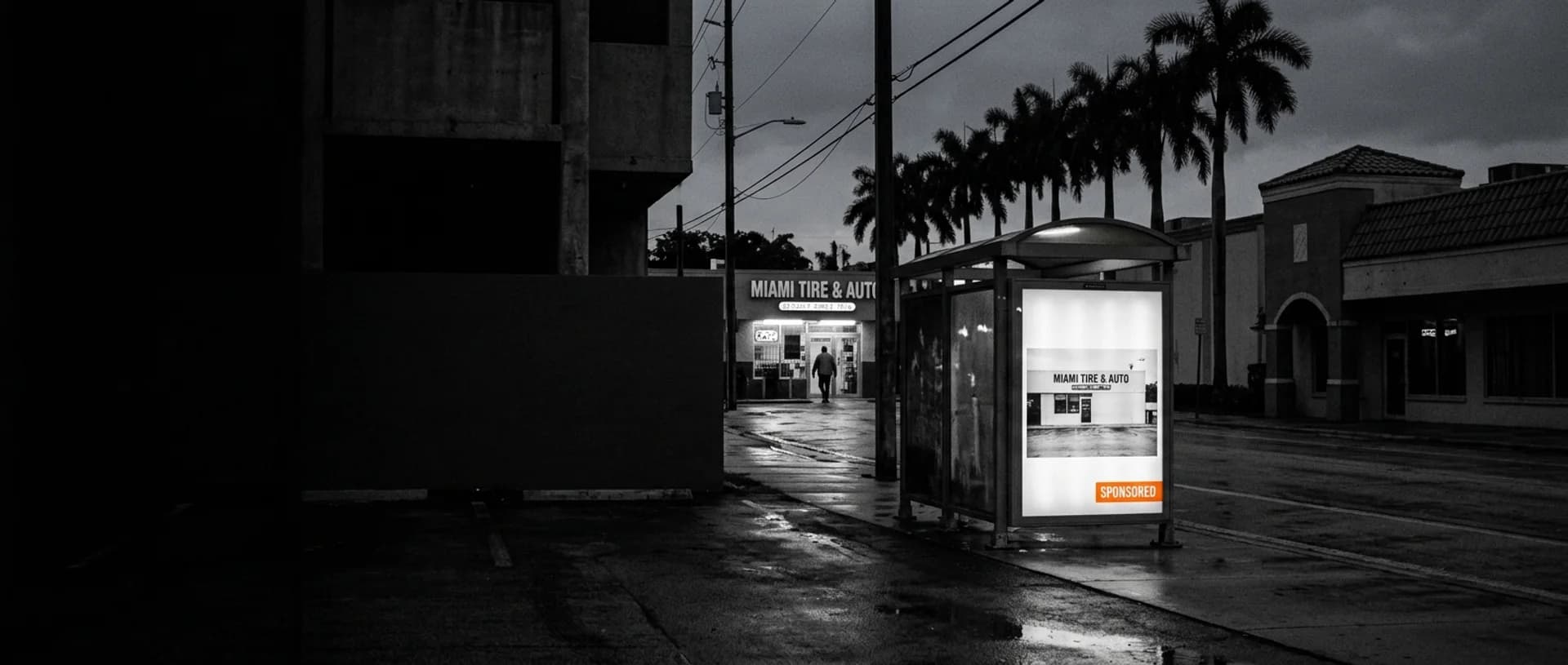 Una marquesina de Miami al atardecer con el panel encendido mostrando un anuncio de Miami Tire & Auto marcado como patrocinado en naranja, y ese mismo taller abierto al otro lado de la calle mojada Una marquesina de Miami al atardecer con el panel encendido mostrando un anuncio de Miami Tire & Auto marcado como patrocinado en naranja, y ese mismo taller abierto al otro lado de la calle mojada